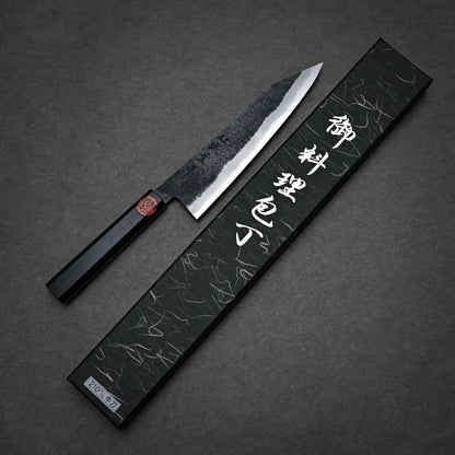 A kurouchi ktip gyuto with a black handle and textured blade rests beside an ornate black box adorned with white Japanese characters, set against a dark background.