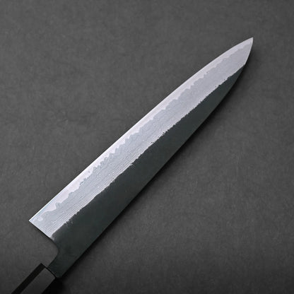 A close-up of a Japanese-style kitchen knife with a shiny, patterned blade and a dark handle, highlighted against a plain dark background. The blade’s intricate design showcases traditional craftsmanship.