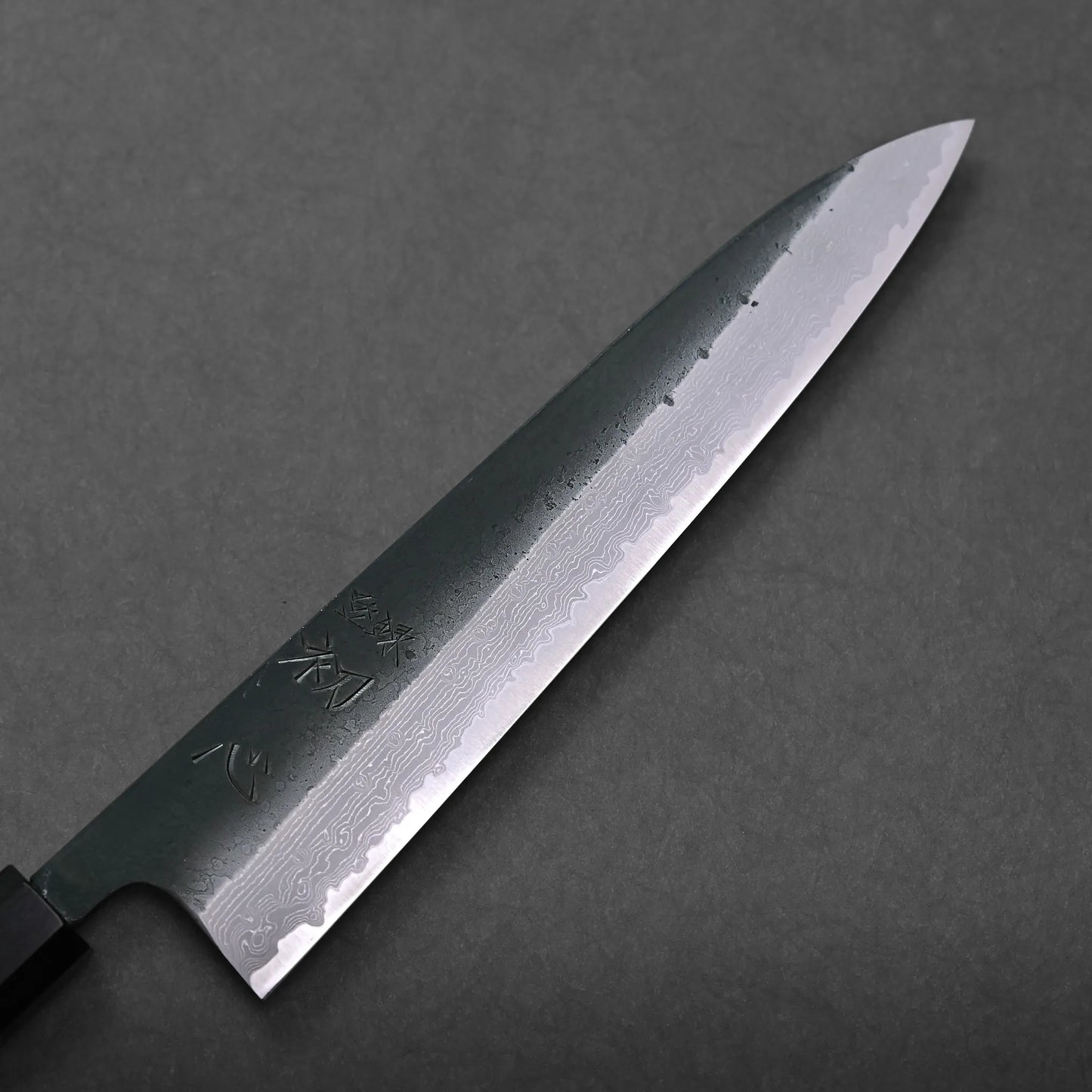 A close-up of a Japanese-style kitchen knife with a damascus steel blade, featuring intricate wave patterns and engraved kanji characters, placed on a dark gray surface for an elegant culinary display.