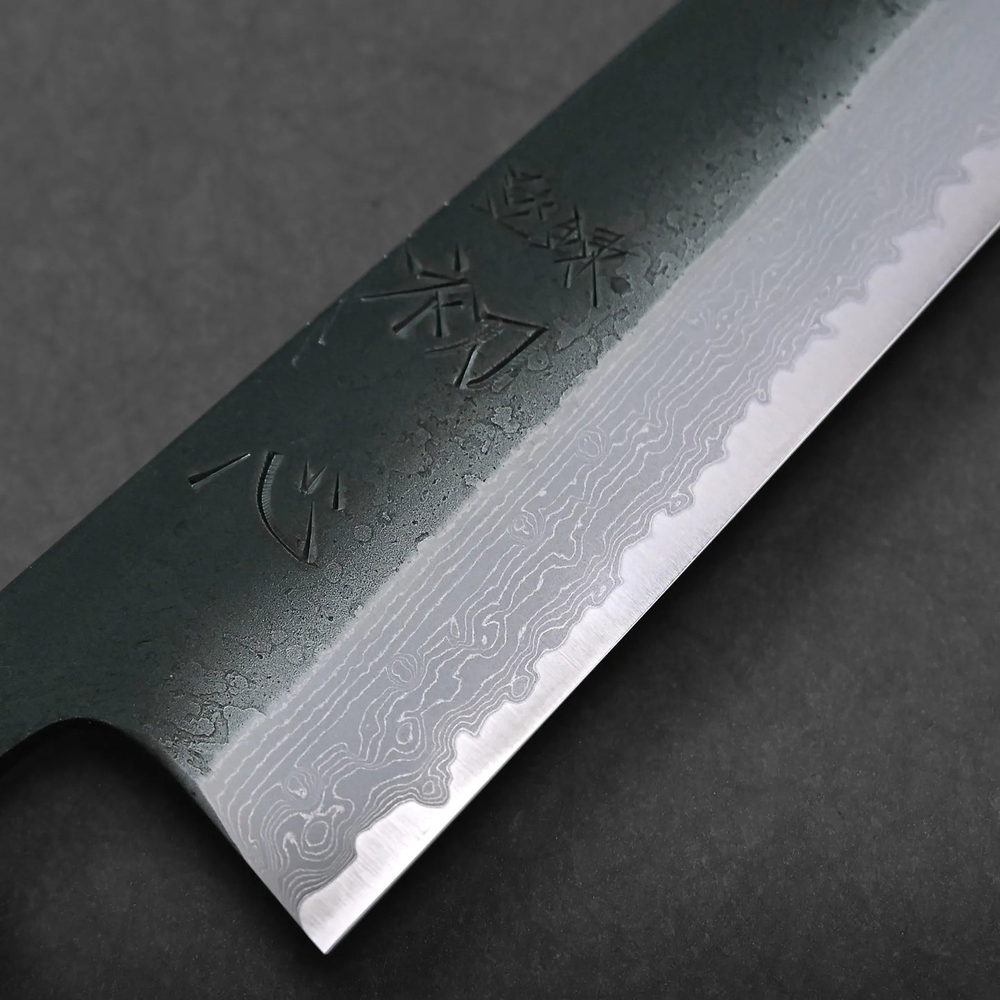 Close-up of a Japanese knife blade featuring striking Damascus steel patterns and engraved Japanese characters on a dark background.