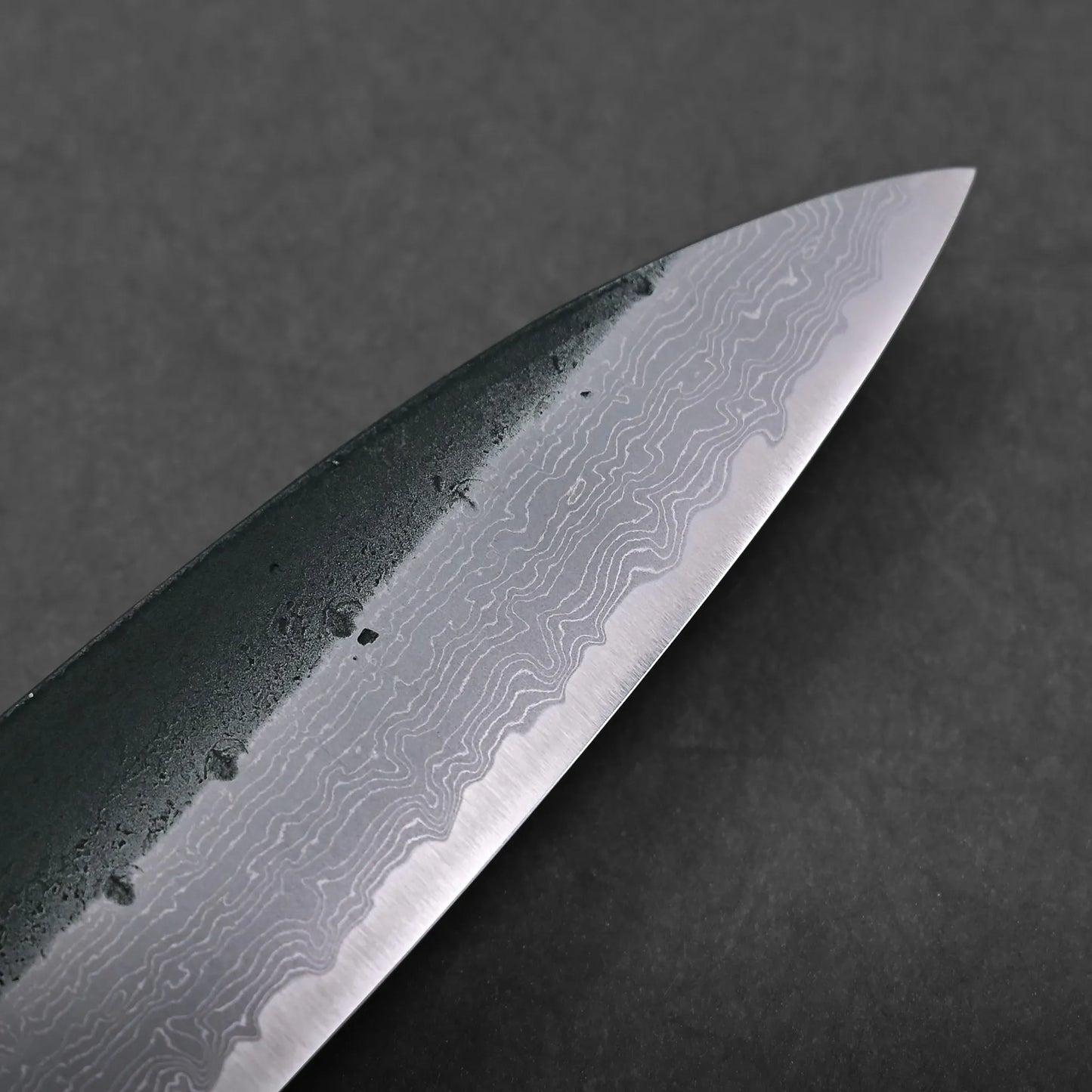 Close-up of a knife blade featuring a distinct wavy Damascus steel pattern, set against a dark background. The blade displays an intricate design and water droplets near the spine.