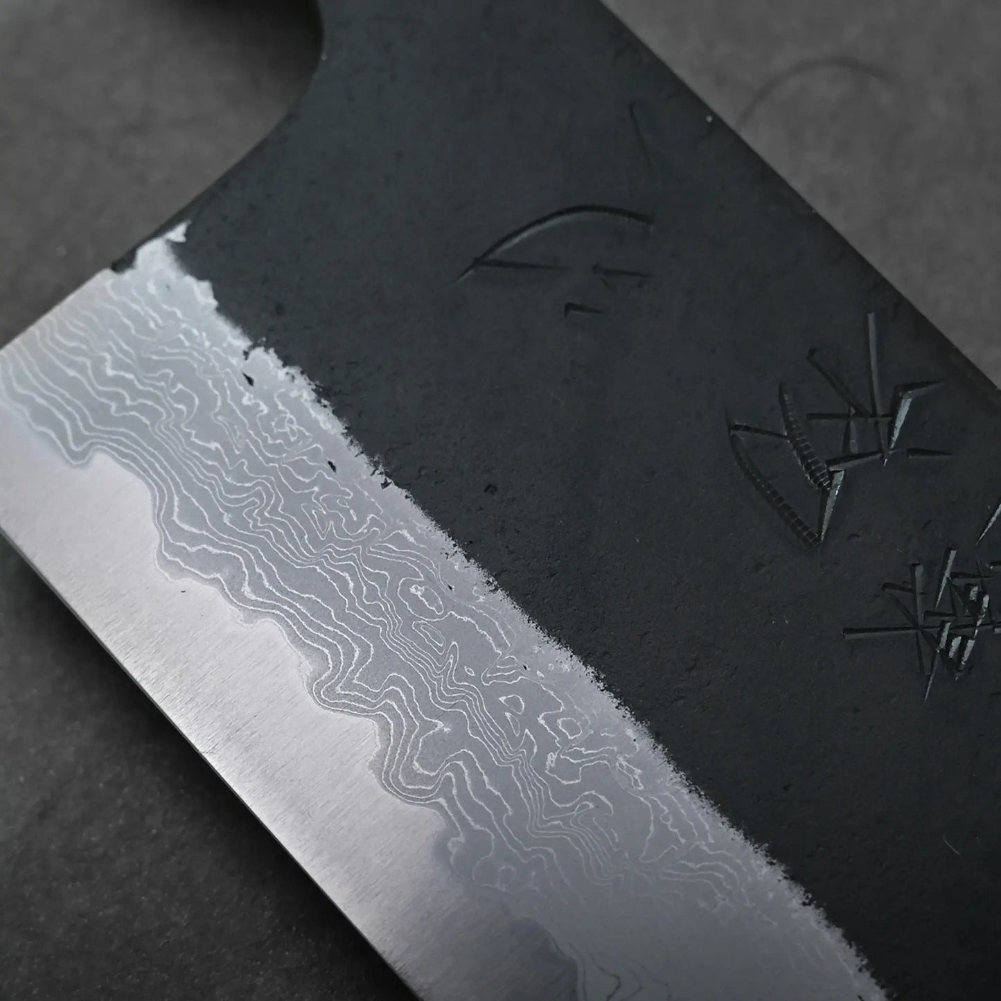 Macro view of a Japanese knife showing kurouchi damascus blade, layered wavy patterns, and engraved symbols.