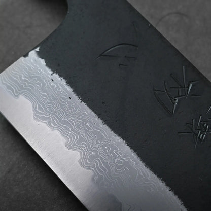 Macro view of a Japanese knife showing kurouchi damascus blade, layered wavy patterns, and engraved symbols.