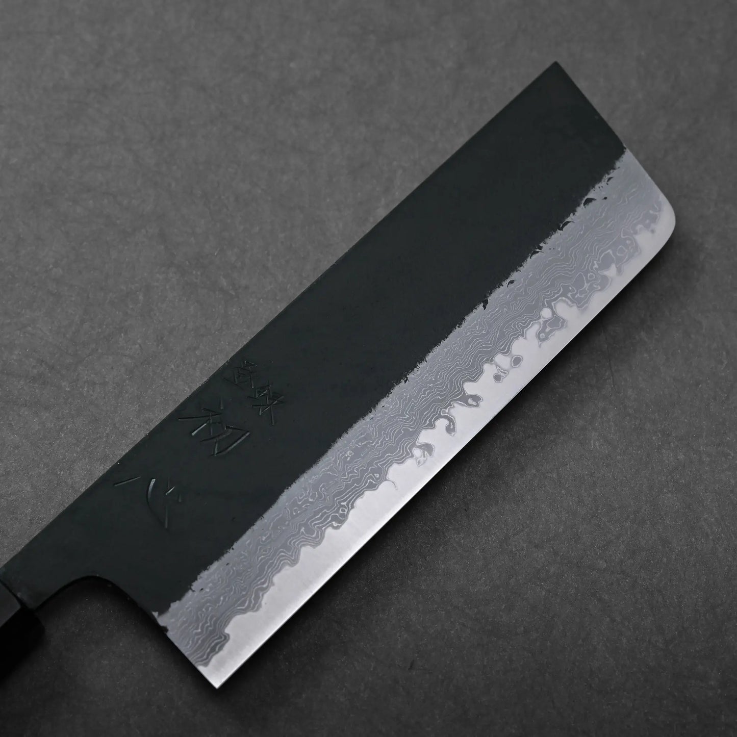 A kurouchi damascus Japanese knife blade with engraved characters shown in close-up, blade shape not fully visible.