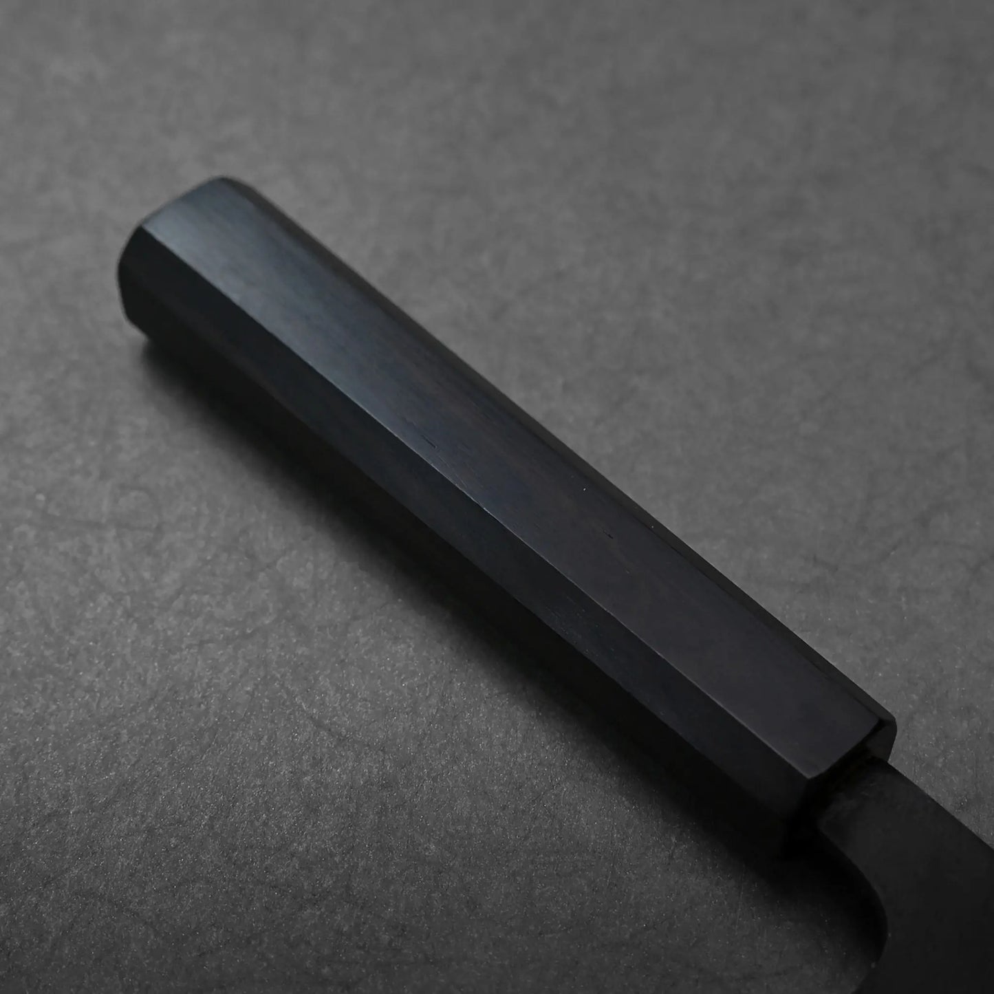 Matte black, hexagonal handle of a hand-forged Japanese knife, close-up with blade not visible in the image.