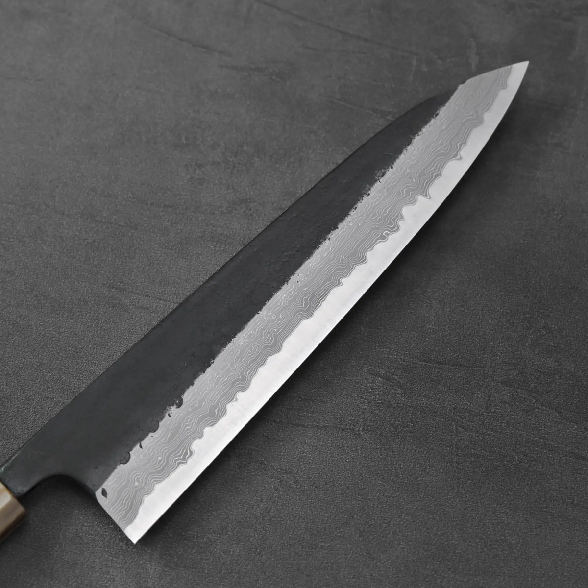 Close-up of a gyuto showing a kurouchi damascus finish and visible cladding lines on the blade.