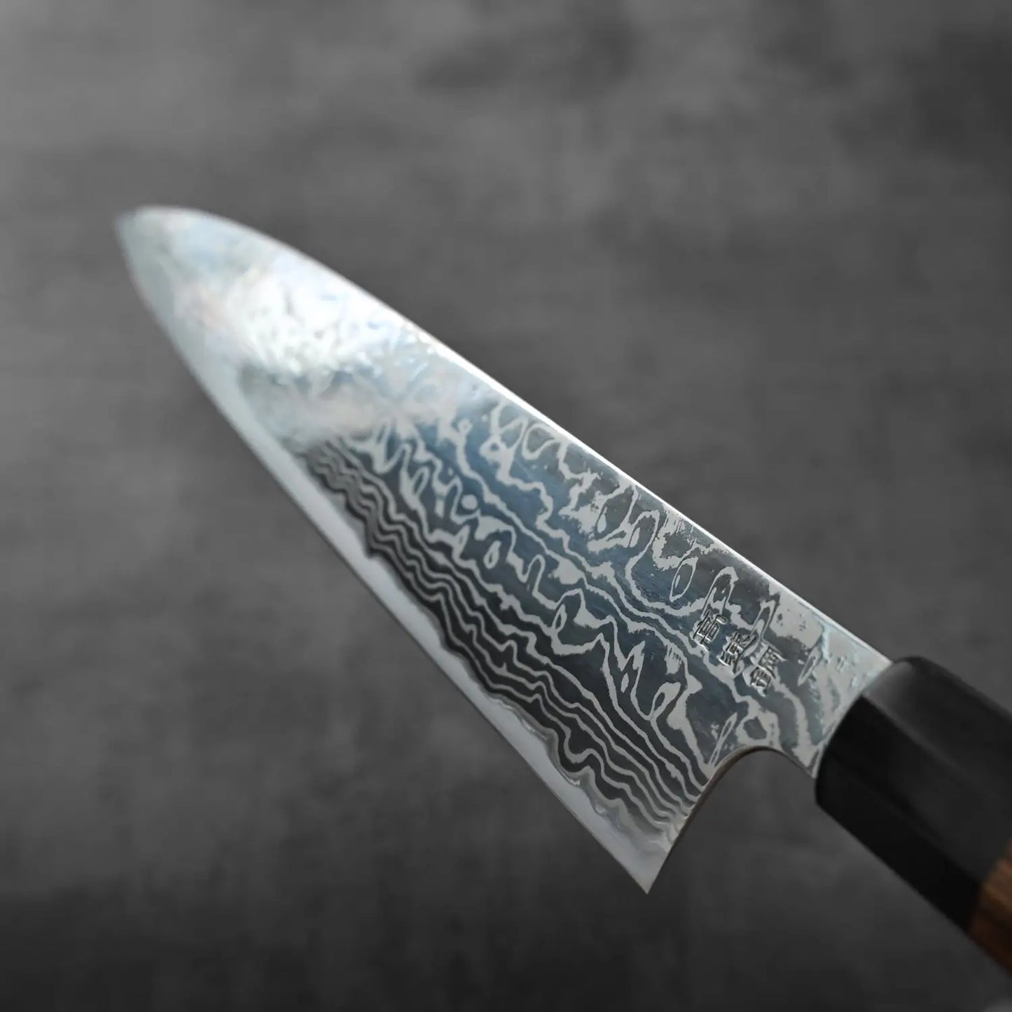 Gyuto with a patterned blade and dark handle, shown in close-up highlighting metal pattern and texture.
