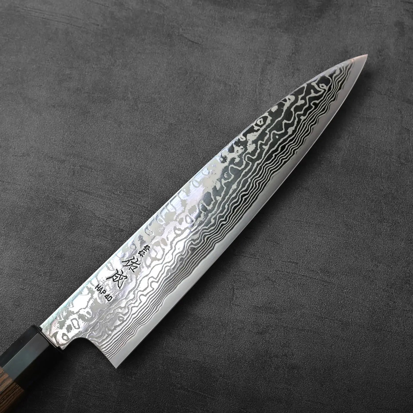 A close-up of a gyuto blade, showing layered Damascus steel pattern and fine detailing on the knife.