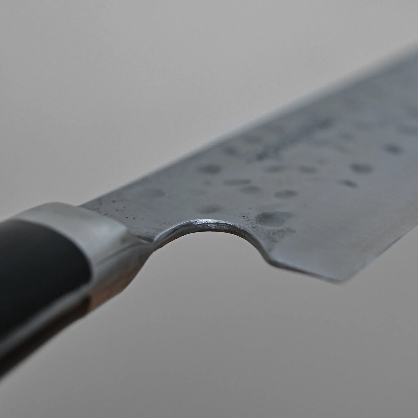 Close-up of the Teruyasu Fujiwara Maboroshi shirogami#1 gyuto 240mm, highlighting its hand-forged hammered blade, pronounced finger notch, partial handle, and a grey background.