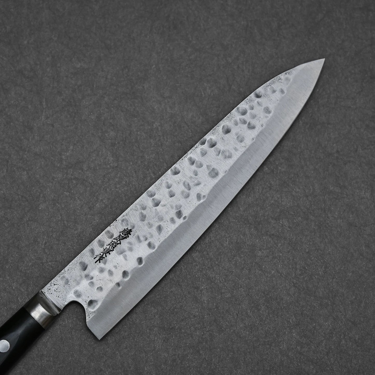 The Teruyasu Fujiwara Maboroshi Shirogami #1 Gyuto 240mm, a hand-forged Japanese knife with a hammered finish and engraved characters near the handle, rests on a dark gray textured surface.