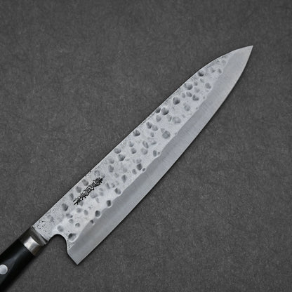The Teruyasu Fujiwara Maboroshi Shirogami #1 Gyuto 240mm, a hand-forged Japanese knife with a hammered finish and engraved characters near the handle, rests on a dark gray textured surface.
