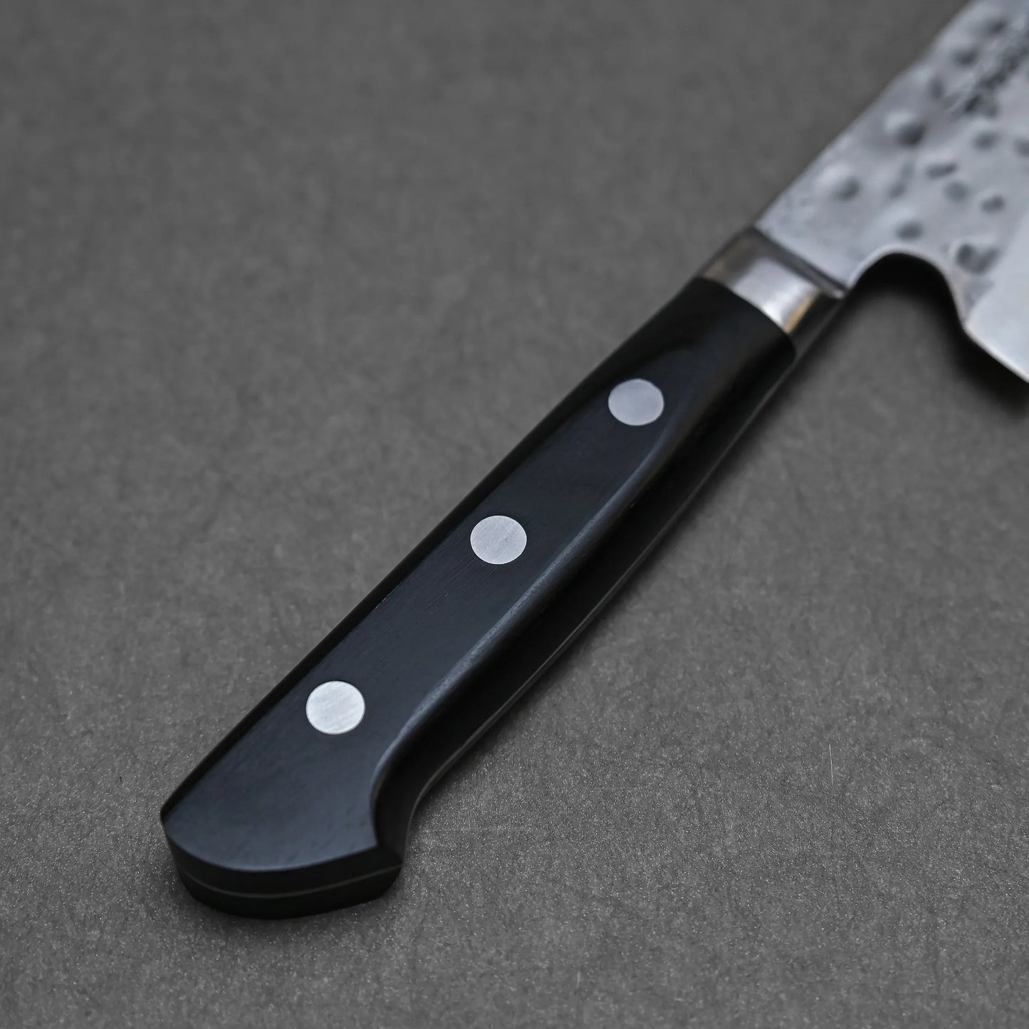 Close-up of a hand-forged Teruyasu Fujiwara Maboroshi shirogami#1 gyuto 240mm handle with three metal rivets, attached to a dark material and resting on a gray surface; part of the blade appears at the top right corner.