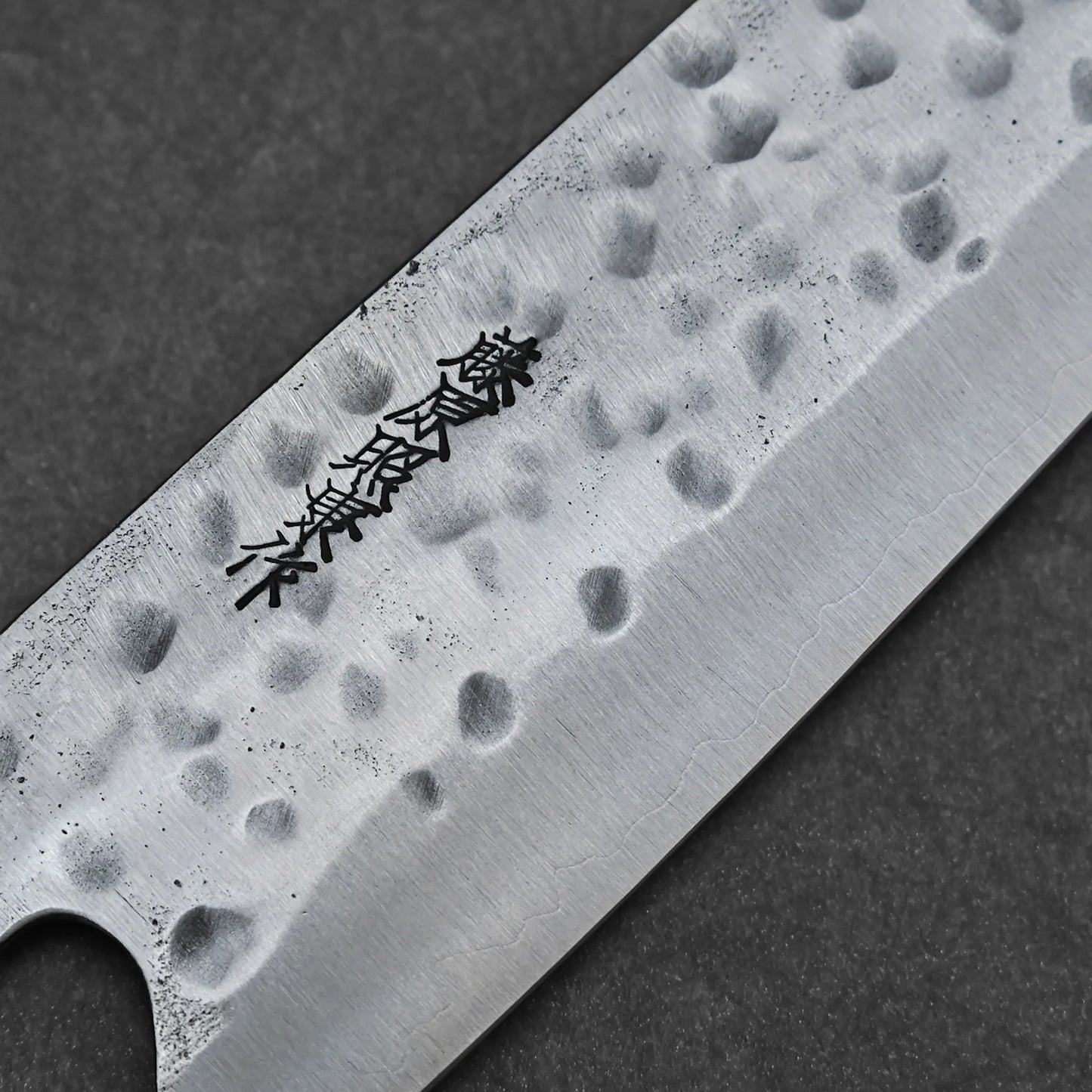 Close-up of the Teruyasu Fujiwara Maboroshi shirogami#1 gyuto 240mm blade, hand-forged with a hammered texture and engraved Japanese characters, set against a dark background.
