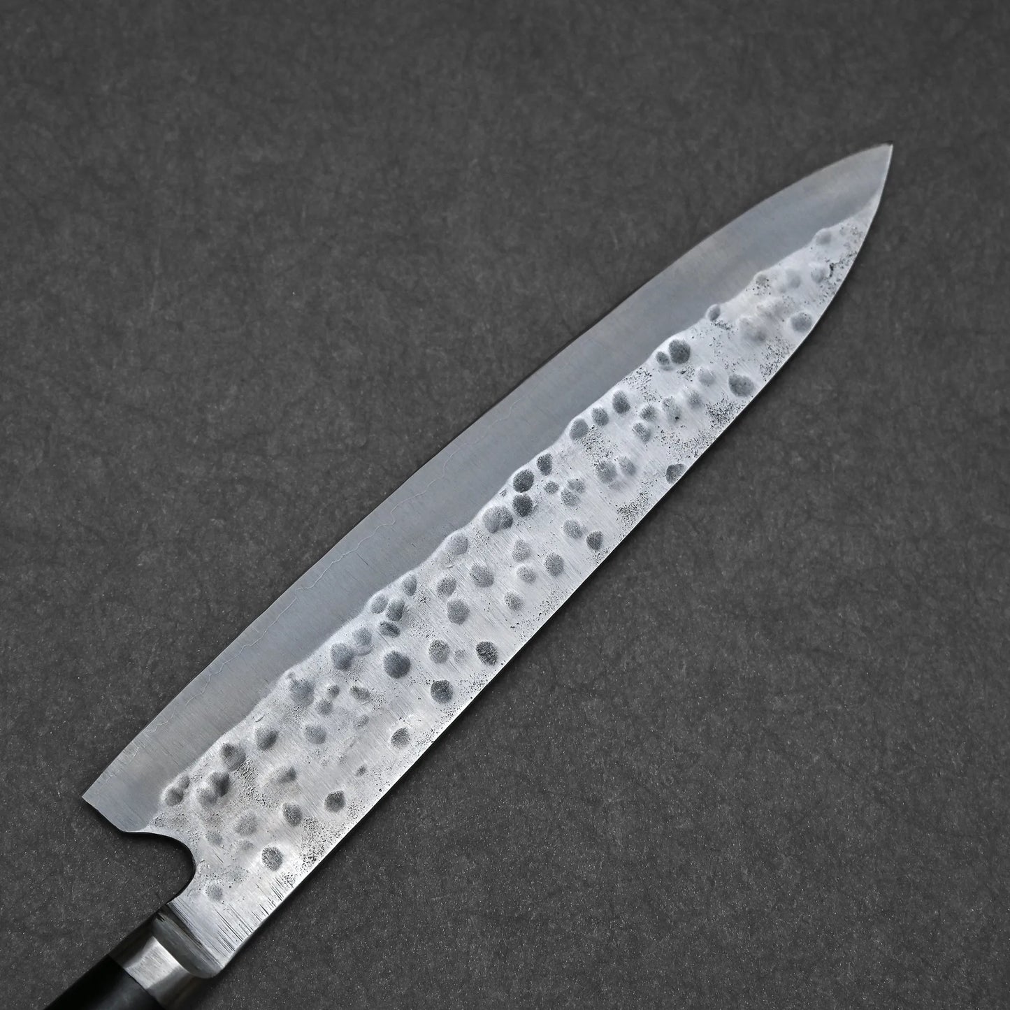 A close-up of the Teruyasu Fujiwara Maboroshi shirogami#1 gyuto 240mm shows its hand-forged, hammered blade with visible patterns and a sharp point, highlighting expert Japanese craftsmanship against a dark, textured background.