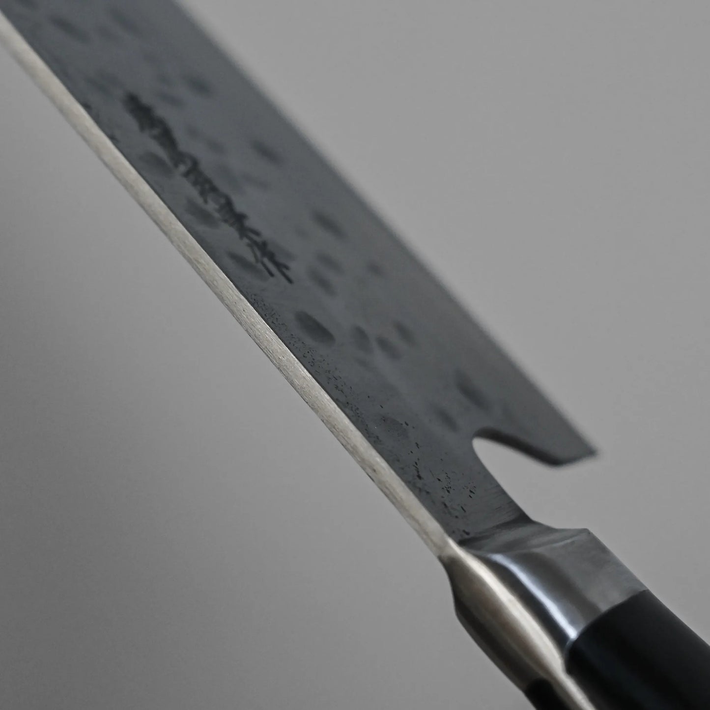 Close-up of a Teruyasu Fujiwara Maboroshi shirogami#1 gyuto 240mm blade with hammered texture and visible edge, shown diagonally on a plain grey background. This hand-forged Japanese knife has a metal bolster and black handle.