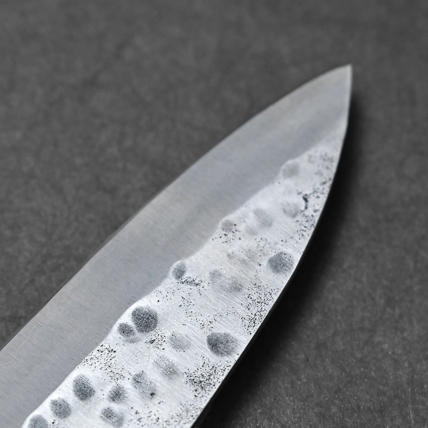 Close-up of the tip of a hand-forged Teruyasu Fujiwara Maboroshi shirogami#1 gyuto 240mm knife, showing its hammered, textured blade against a dark background. The edge looks exceptionally sharp and polished.