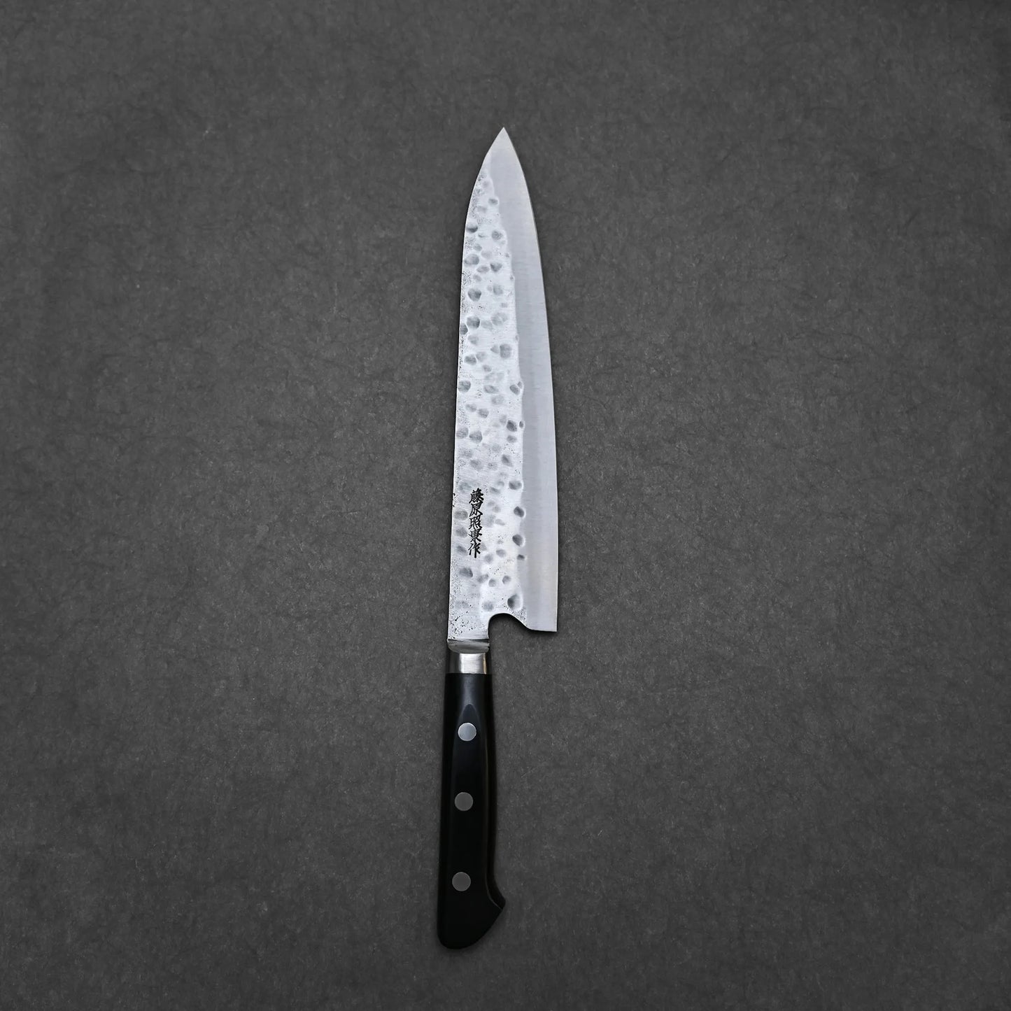 The Teruyasu Fujiwara Maboroshi shirogami#1 gyuto 240mm, a hand-forged Japanese chef knife with a hammered finish and black handle, rests on a gray textured surface.