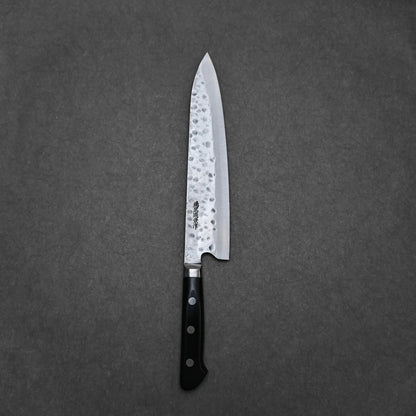 The Teruyasu Fujiwara Maboroshi shirogami#1 gyuto 240mm, a hand-forged Japanese chef knife with a hammered finish and black handle, rests on a gray textured surface.
