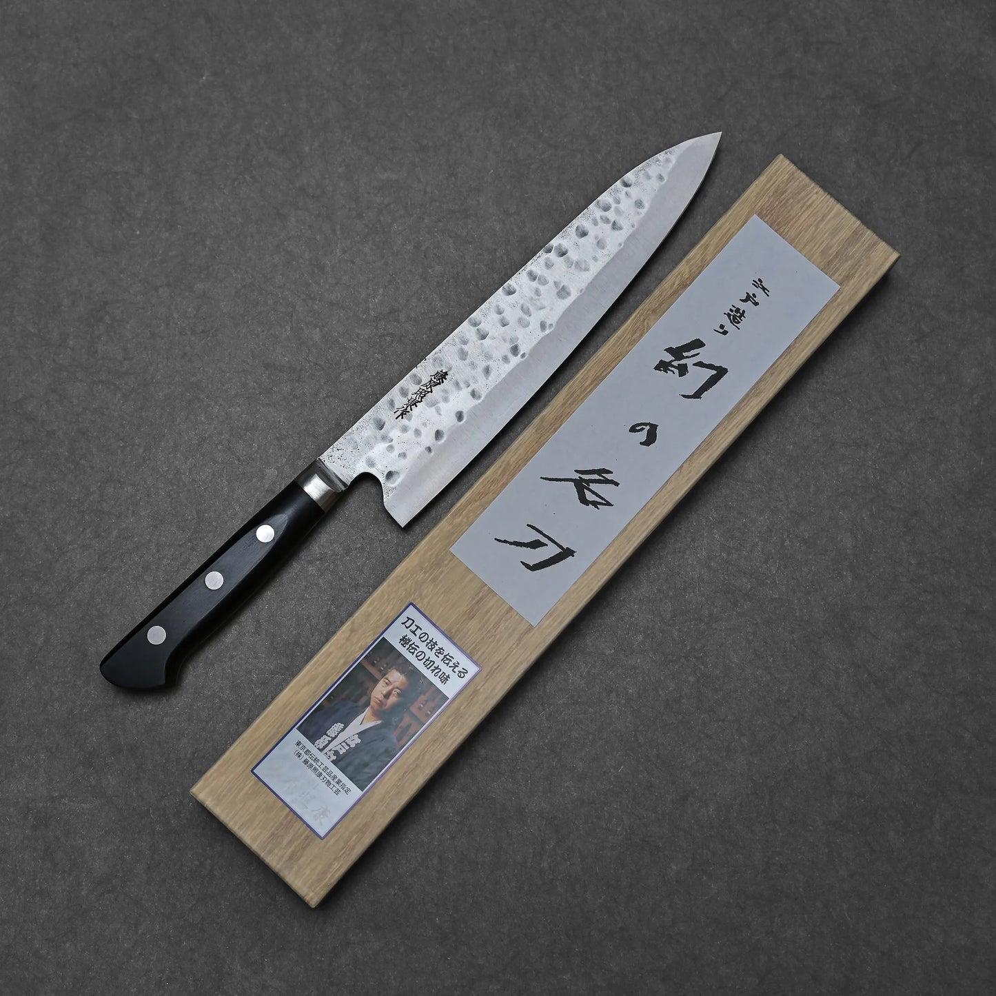 The Teruyasu Fujiwara Maboroshi shirogami#1 gyuto 240mm, featuring a hand-forged hammered blade and black handle, rests beside a wooden presentation box with Japanese calligraphy and a photo label on a dark gray surface.