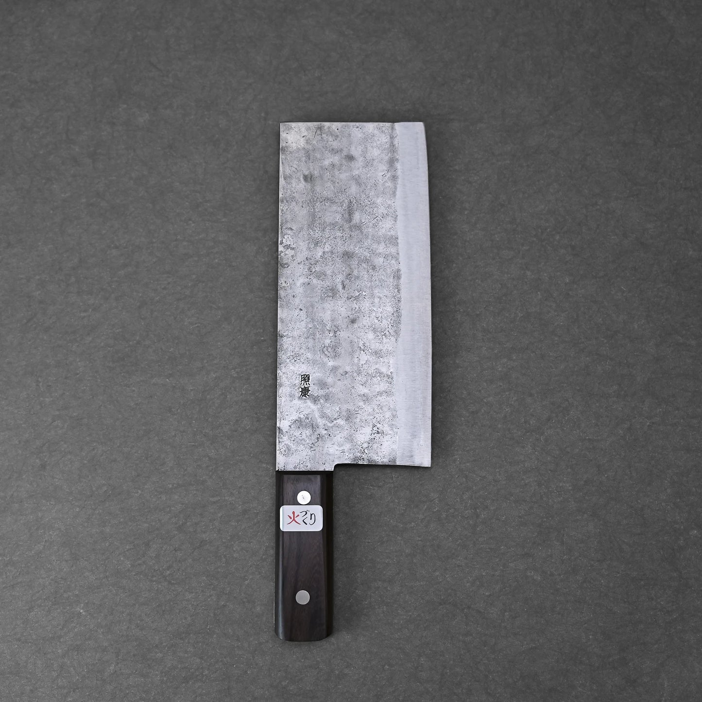 A Teruyasu Fujiwara nashiji shirogami#1 chuka bocho 210x90mm, featuring a dark wooden handle and rectangular, textured carbon steel blade, rests on a flat, dark gray surface.