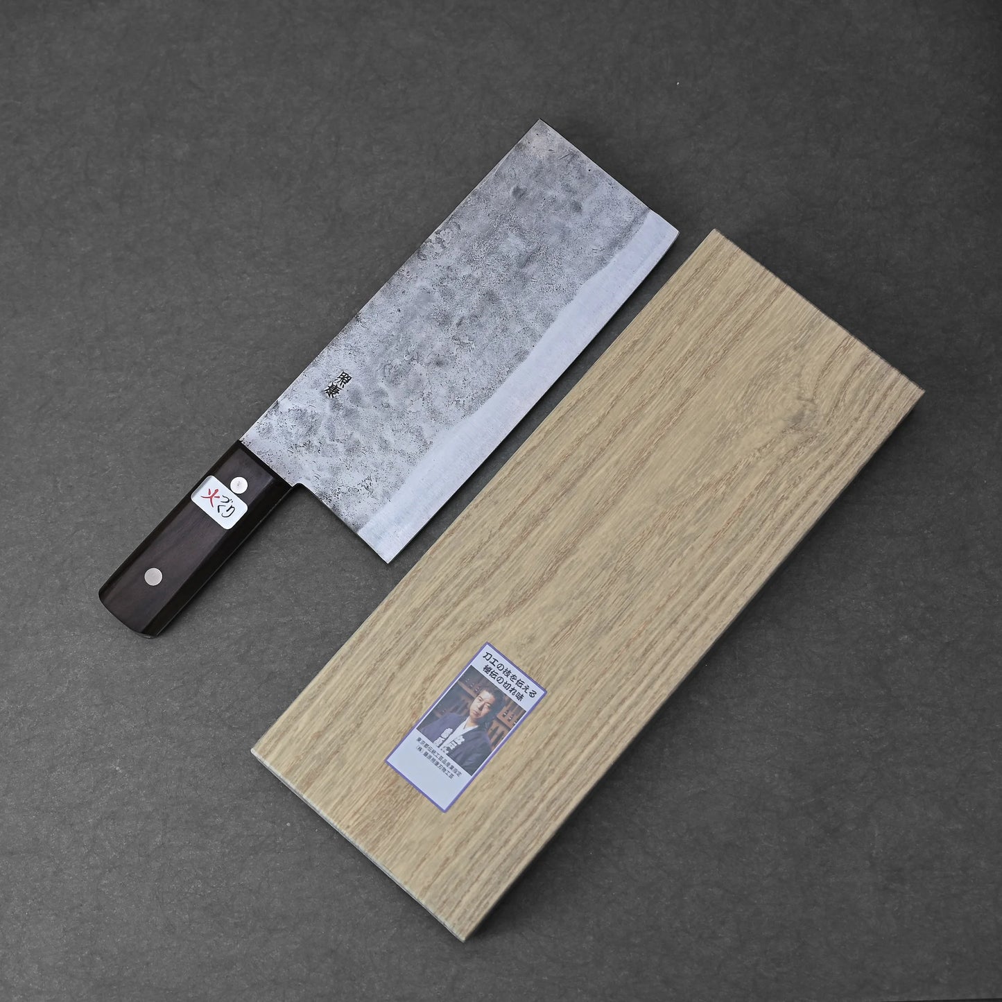 A Teruyasu Fujiwara nashiji shirogami#1 chuka bocho 210x90mm with a dark handle and carbon steel blade rests beside a light wooden box featuring a small label with text and a photo, all on a dark surface.