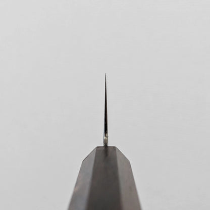 A vertical close-up shot of the choil of a gyuto knife, seen from directly below. The polished ginsan stainless steel blade tapers sharply upwards to a fine point, emerging from the dark mono octagonal ebony handle, which occupies the lower portion of the frame.