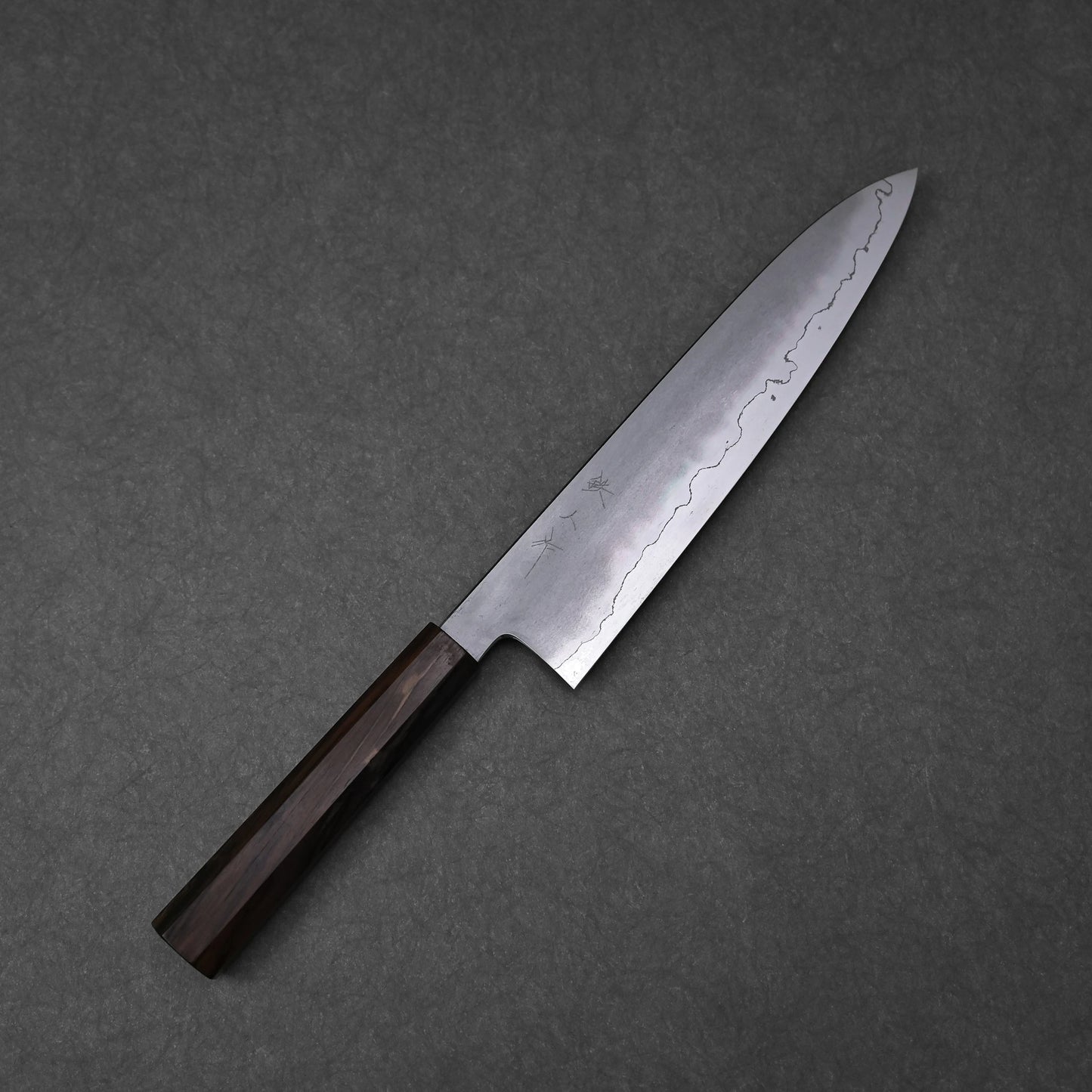 A full view of a gyuto knife, positioned diagonally on a textured grey surface. The ginsan stainless steel blade features a kasumi finish with a wavy boundary between the polished edge and the frosted upper surface. Subtle kanji engravings are visible on the blade, which is fitted with a dark, mono octagonal ebony handle.