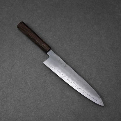 A full, overhead view of a gyuto knife, positioned diagonally on a textured grey surface. The ginsan stainless steel blade displays a kasumi finish with a prominent wavy border and subtle kanji engravings on its upper surface. The knife is fitted with a dark, mono octagonal ebony handle.
