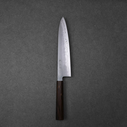 A full, overhead view of a gyuto knife, positioned vertically on a dark grey surface. The ginsan stainless steel blade features a kasumi finish with a wavy border and kanji engravings on the upper surface. The knife has a dark, mono octagonal ebony handle at the bottom.