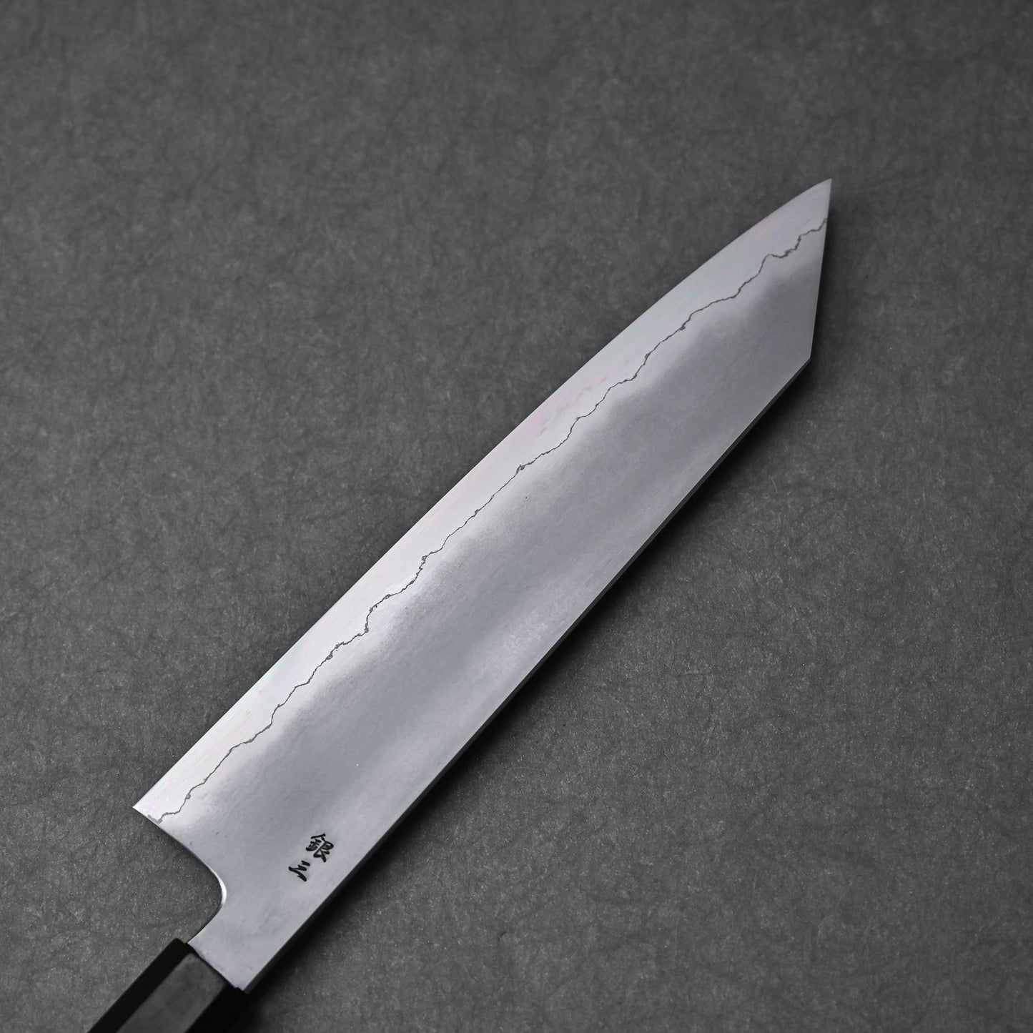 A full view of the reverse side of a kiritsuke gyuto knife blade. The ginsan stainless steel blade displays a clear kasumi finish with a distinct, wavy border. Kanji engravings are visible near the heel of the blade.