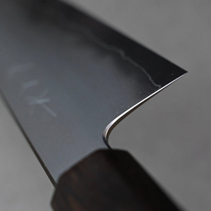 A close-up, angled view of the choil of a kiritsuke gyuto knife, where the blade meets the handle. The clean finish of the ginsan stainless steel is visible, curving smoothly into the polished edge. The dark ebony handle is partially visible at the bottom.