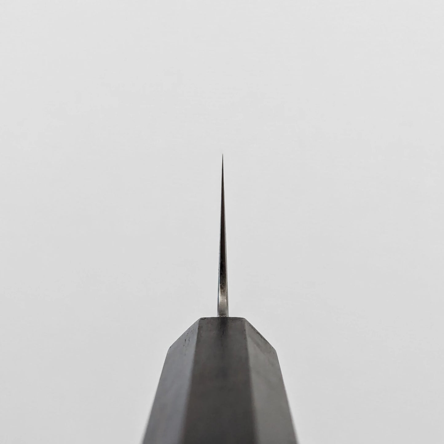 A vertical close-up shot of the choil of a kiritsuke gyuto knife, seen from directly below. The polished ginsan stainless steel blade tapers sharply upwards to a fine point, emerging from the dark octagonal ebony handle, which occupies the lower portion of the frame.