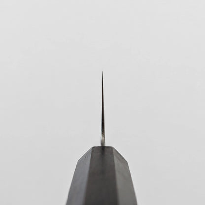 A vertical close-up shot of the choil of a kiritsuke gyuto knife, seen from directly below. The polished ginsan stainless steel blade tapers sharply upwards to a fine point, emerging from the dark octagonal ebony handle, which occupies the lower portion of the frame.