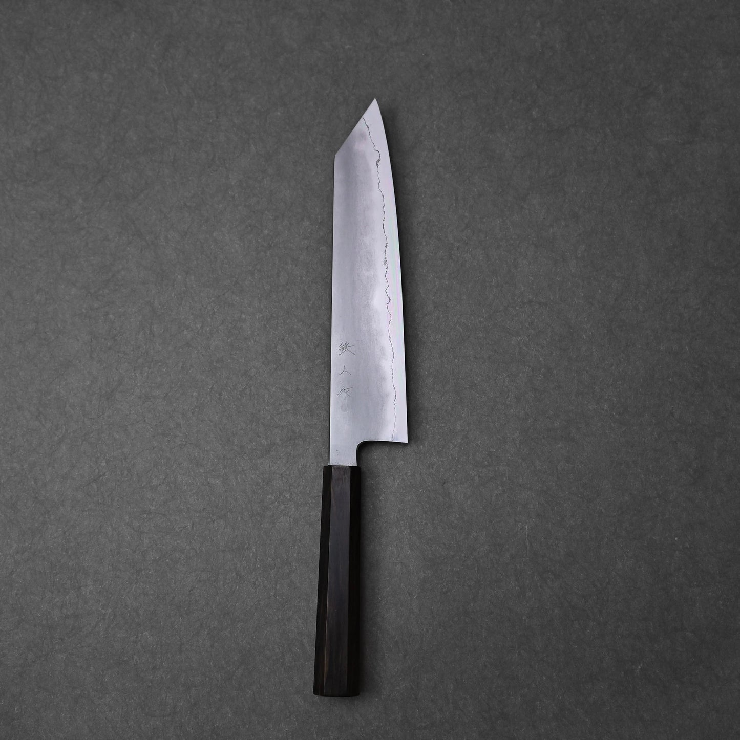 A full, overhead view of a kiritsuke gyuto knife, positioned vertically on a dark grey surface. The ginsan stainless steel blade features a kasumi finish with a wavy border and kanji engravings on the upper surface, tapering to a sharply angled tip. The knife has a dark, octagonal ebony handle at the bottom.
