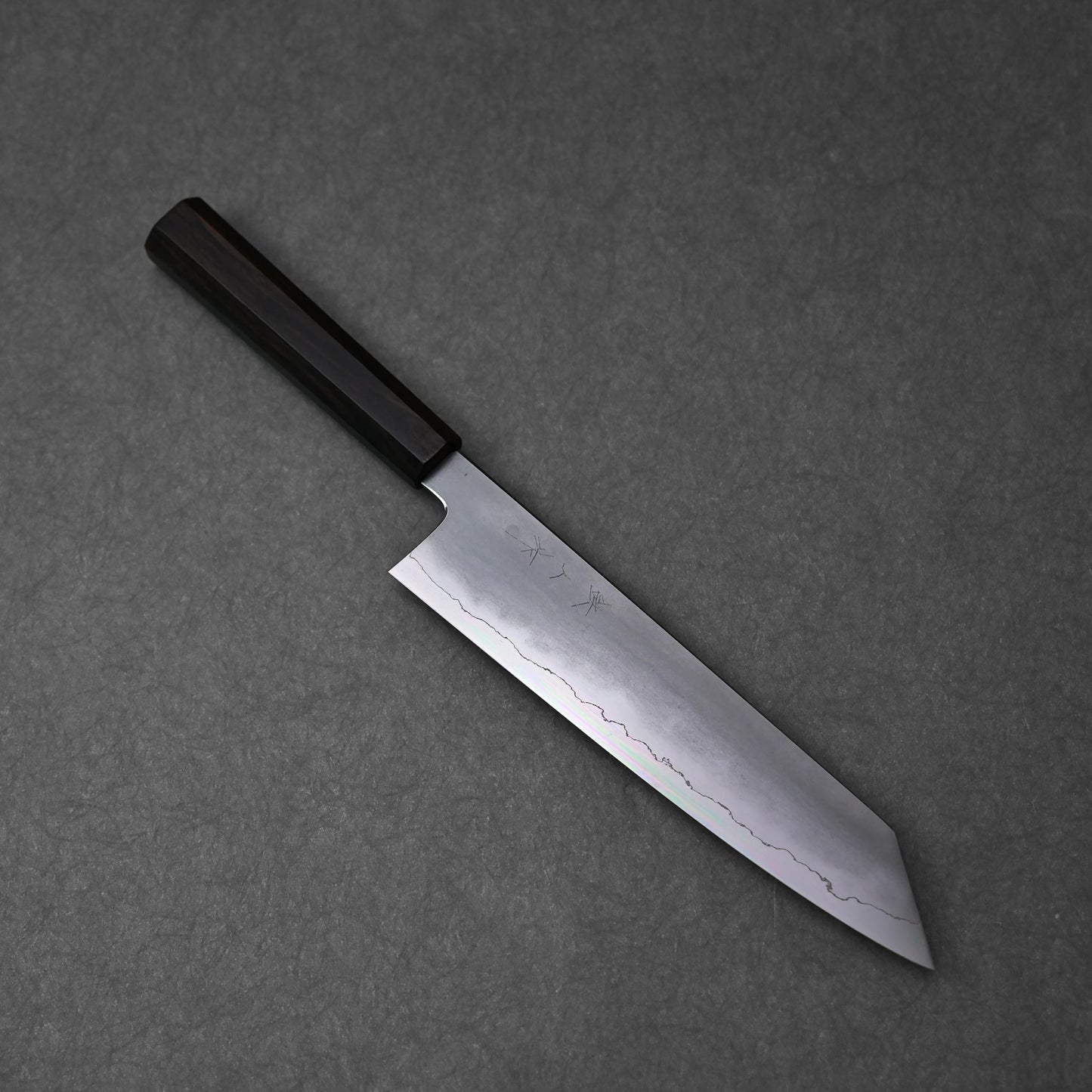 A full, overhead view of a kiritsuke gyuto knife, positioned diagonally on a dark grey surface. The ginsan stainless steel blade features a kasumi finish with a prominent wavy border, and subtle kanji engravings are visible on its upper surface. The knife is fitted with a dark, octagonal ebony handle.