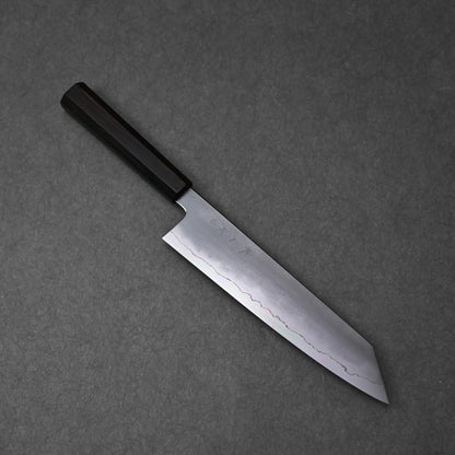 A full, overhead view of a kiritsuke gyuto knife, positioned diagonally on a dark grey surface. The ginsan stainless steel blade features a kasumi finish with a prominent wavy border, and subtle kanji engravings are visible on its upper surface. The knife is fitted with a dark, octagonal ebony handle.