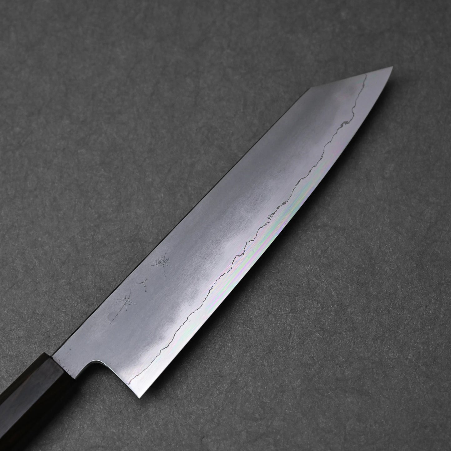 A detailed close-up of a kiritsuke gyuto knife blade, highlighting its ginsan stainless steel construction and kasumi finish. The image shows the subtle, cloud-like texture on the upper portion of the blade transitioning to a reflective, polished cutting edge. Light subtly catches the different finishes along the blade's length.
