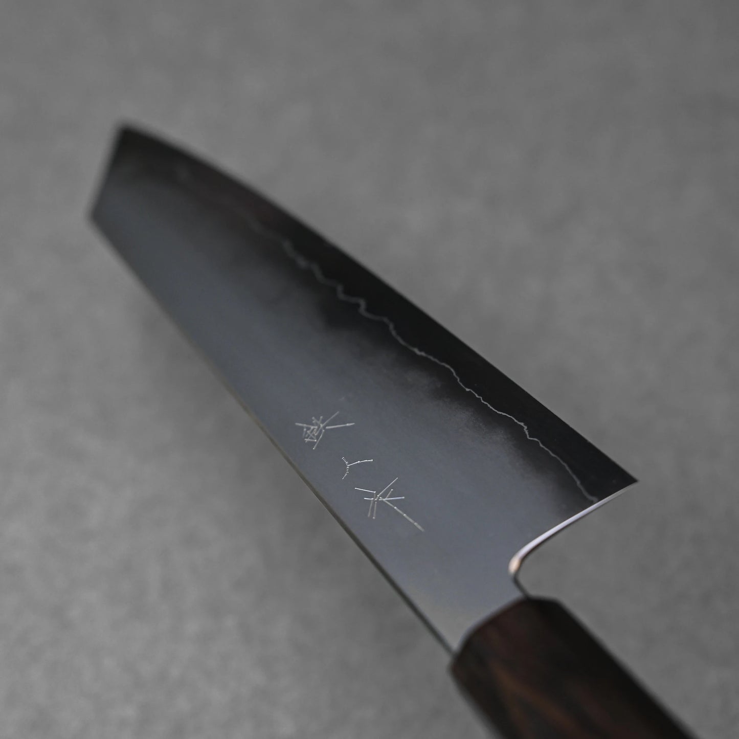 A close-up, angled view of the ginsan stainless steel blade of a kiritsuke gyuto knife. The kasumi finish is visible, showing a darker upper section and a brighter, polished edge. Kanji characters are subtly engraved on the upper part of the blade.