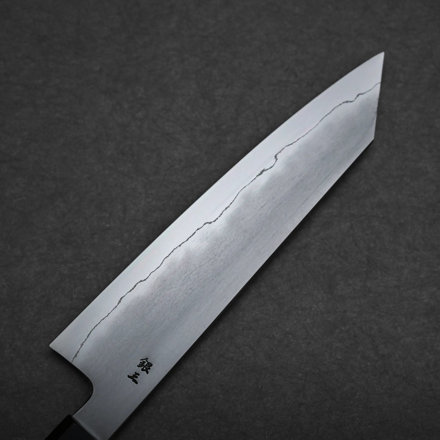A close-up of a kiritsuke gyuto blade with a visible wavy hamon line and small black kanji characters near the handle, set against a dark textured background.