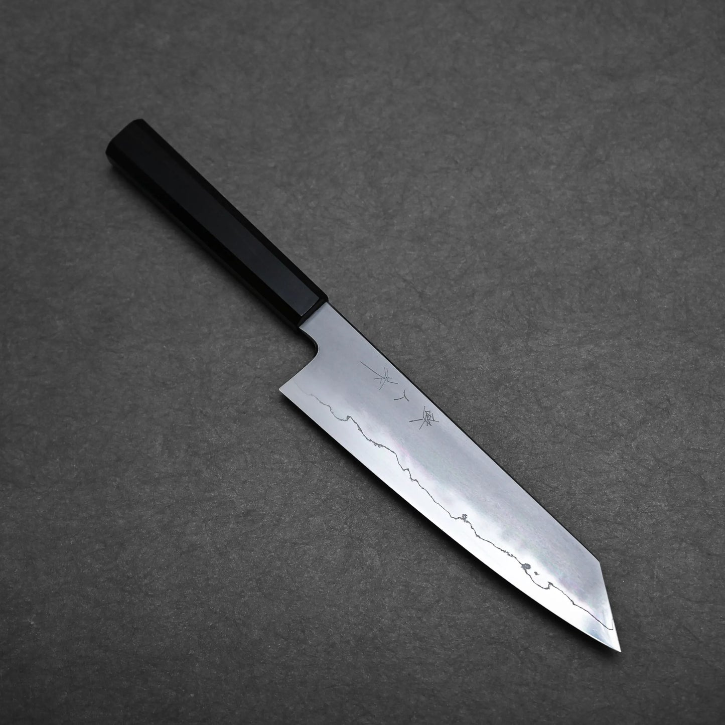 A Japanese-style kiritsuke chef knife with a black handle and a straight-edged blade featuring a distinct wavy pattern, placed on a dark gray textured surface.