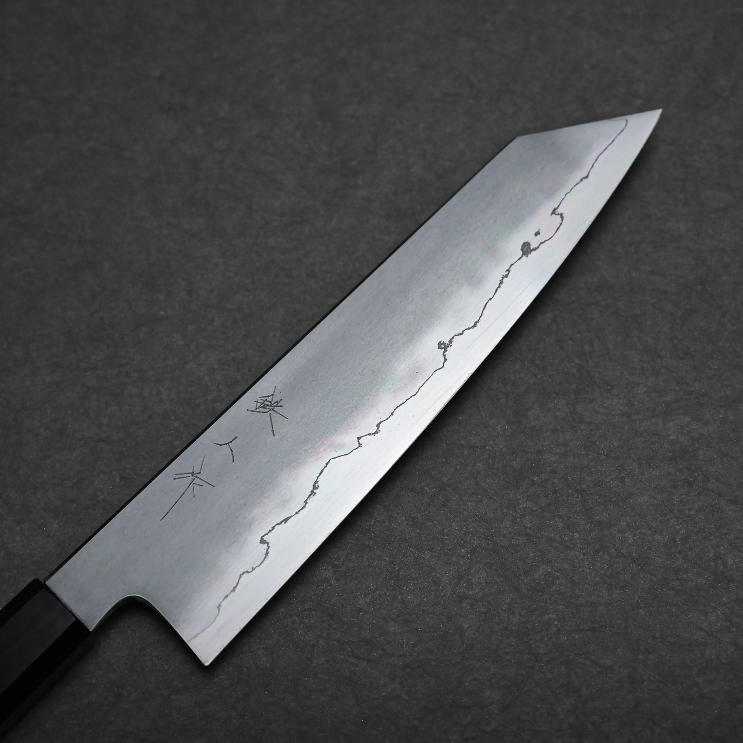 A close-up of a kiritsuke Japanese chef knife with a straight edge and pointed tip, showing a visible wavy line pattern on the blade and Japanese characters etched near the handle, placed on a dark textured surface.