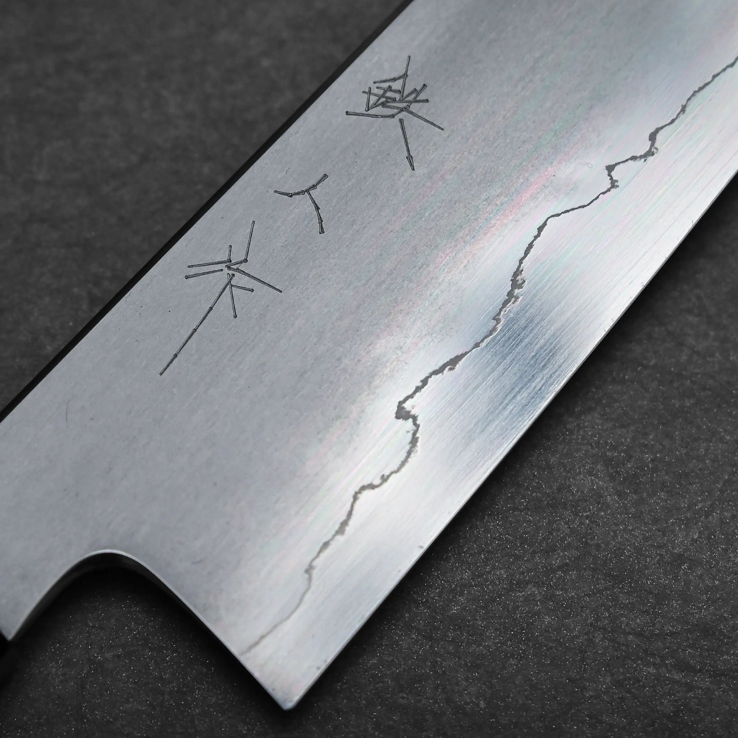 A close-up of a kiritsuke Japanese chef knife blade shows engraved characters and a visible wavy line pattern along the edge, set against a dark textured background.