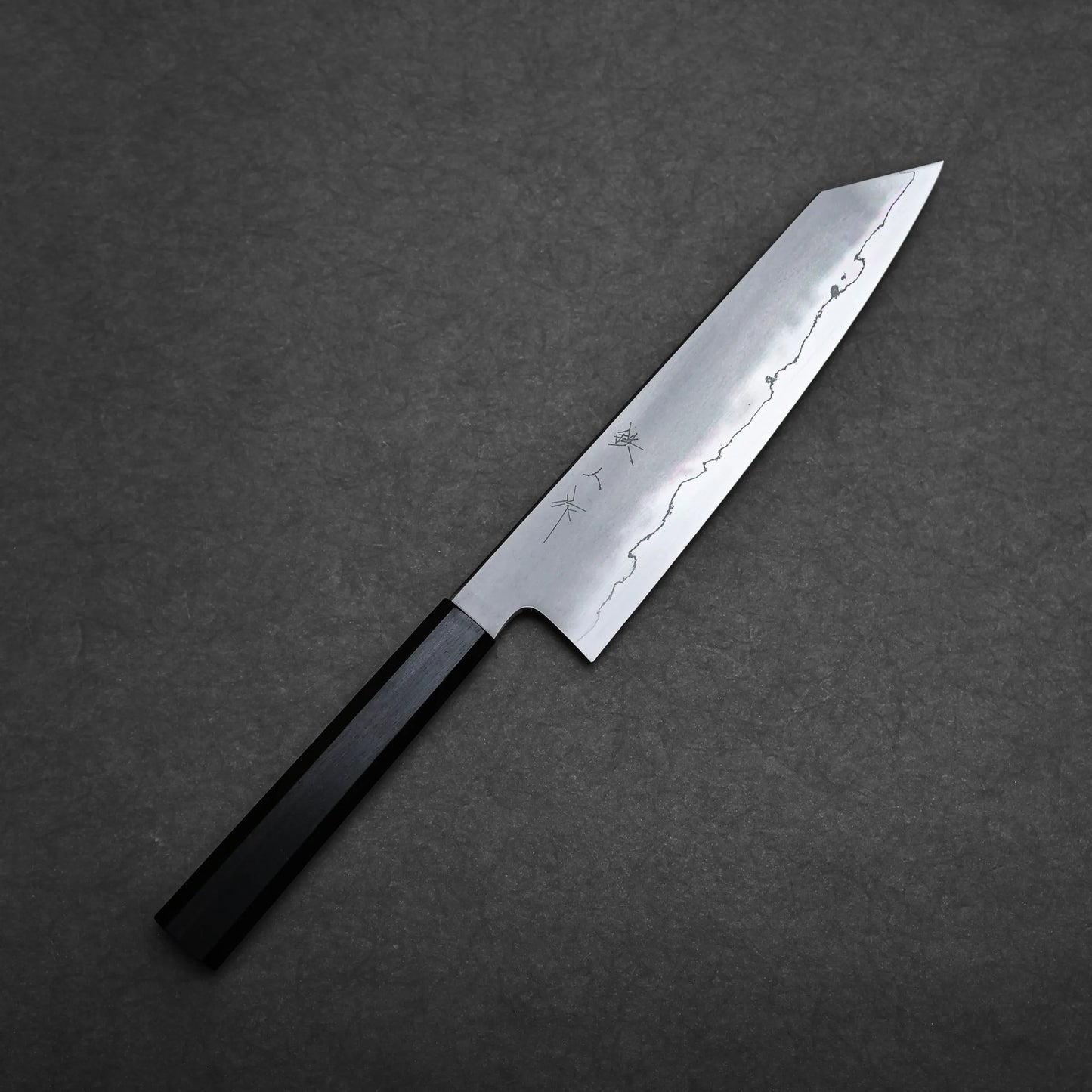 A Tetsujin kasumi ginsan kiritsuke gyuto 210mm with a black handle and a sharp, silver blade featuring subtle decorative patterns, placed on a dark gray textured surface.