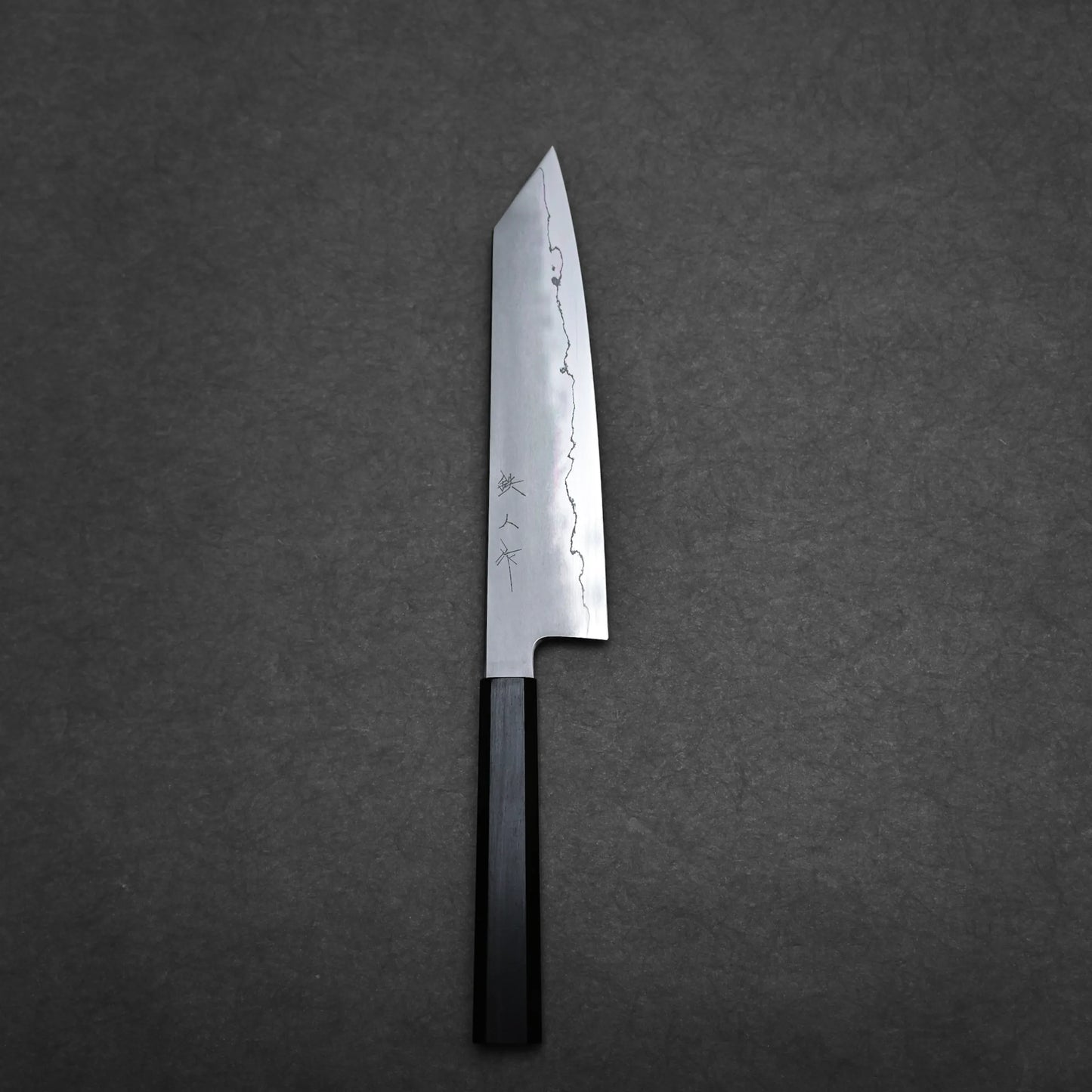 A single kiritsuke Japanese chef knife with a dark handle and a polished, straight-edged blade engraved with Japanese characters, placed on a dark, textured background.