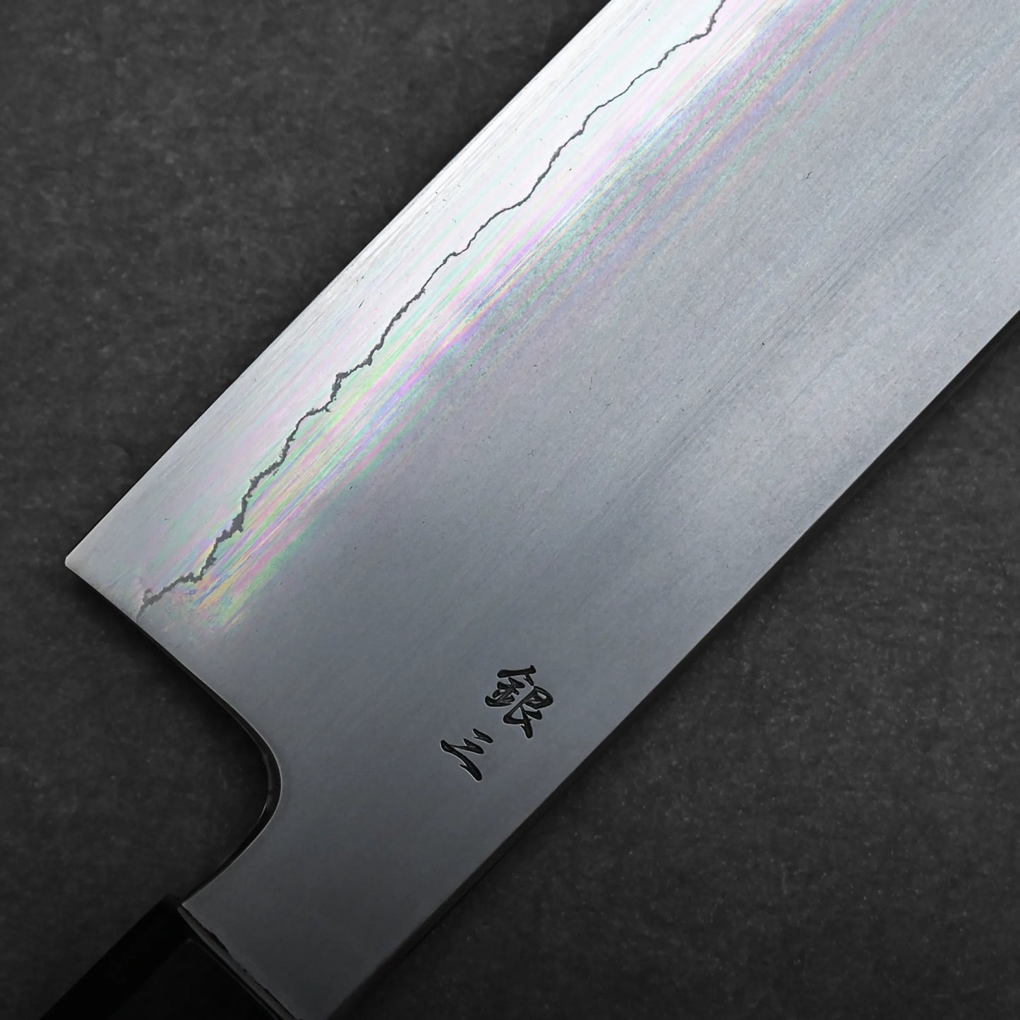Close-up of a Japanese vegetable knife blade, showing a wavy hamon line near the edge, iridescent reflections on the metal, and Japanese characters engraved near the handle, set against a dark textured background.