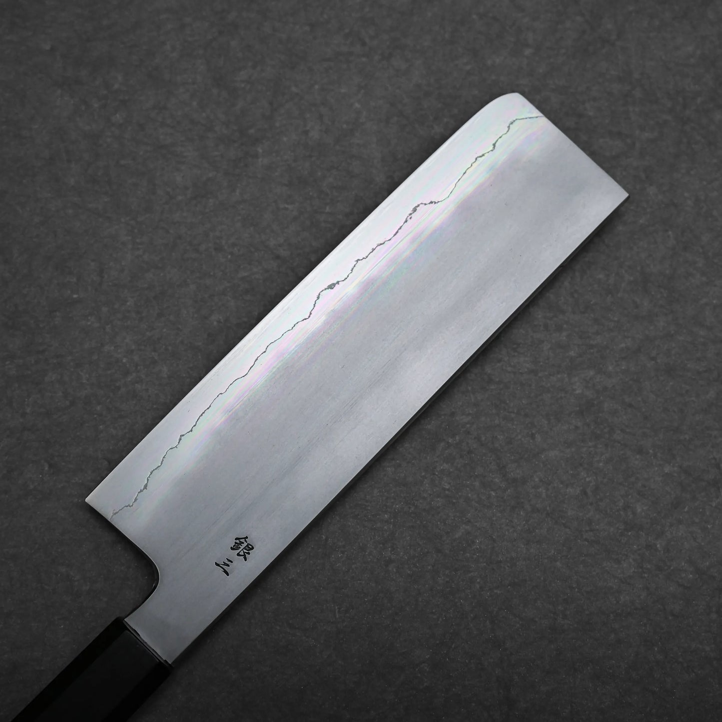 A rectangular Japanese vegetable knife, known as a nakiri knife, with a straight edge and wavy hamon line rests on a dark textured surface. The handle is partially visible.