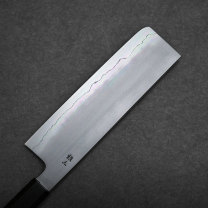 A rectangular Japanese vegetable knife, known as a nakiri knife, with a straight edge and wavy hamon line rests on a dark textured surface. The handle is partially visible.
