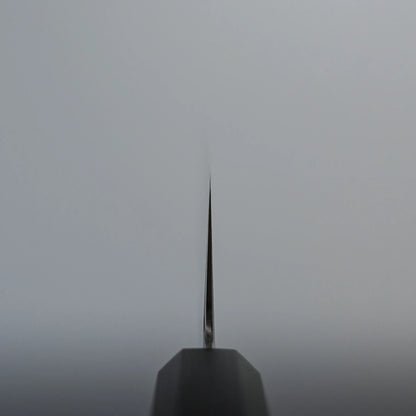 Choil shot of a nakiri knife blade with the sharp edge facing toward the viewer, making the thin metal appear as a vertical line against a plain, light-colored background.