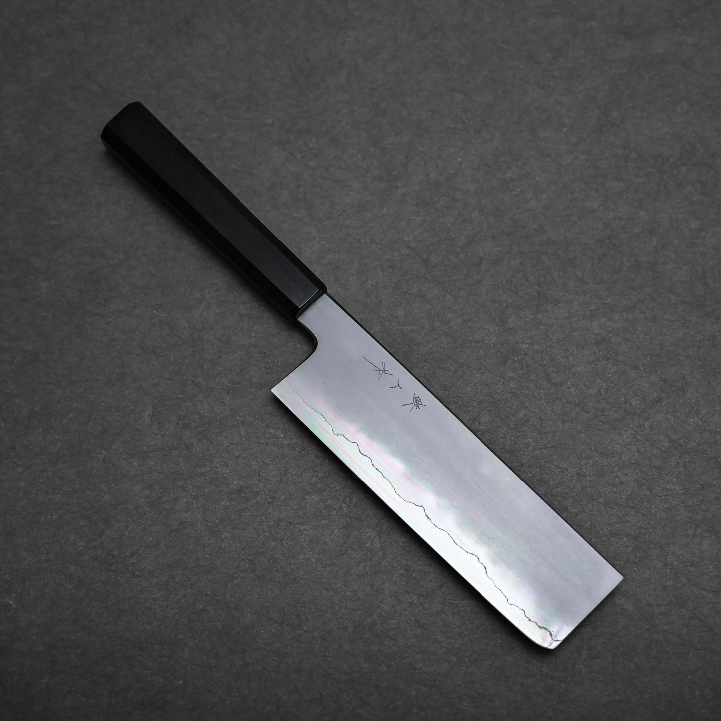 A Japanese vegetable knife, or nakiri knife, with a straight-edged, rectangular blade and a dark handle rests on a dark, textured surface. Japanese characters are engraved on the blade.