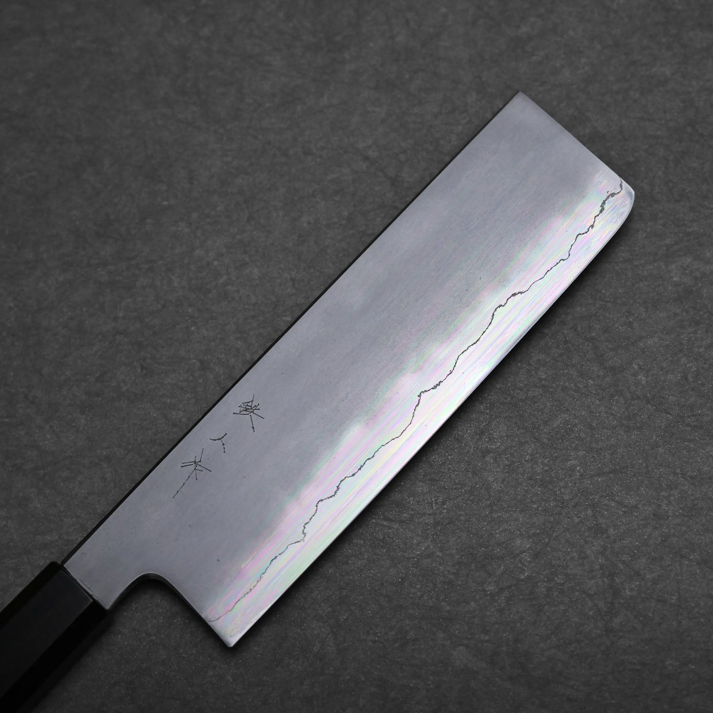 A Japanese vegetable knife, or nakiri knife, with a rectangular blade featuring a wavy pattern and engraved Japanese characters, lies on a dark textured surface.